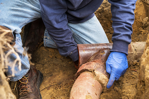 Plumbing Solutions LLC technician repairing a damaged underground sewer pipe inside an excavation trench.