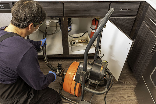 Plumbing Solutions LLC plumber clearing a clogged kitchen sink using an electric drain snake under the cabinet.