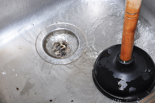 Plumbing Solutions LLC technician unclogging a stainless steel kitchen sink with a plunger as water drains.