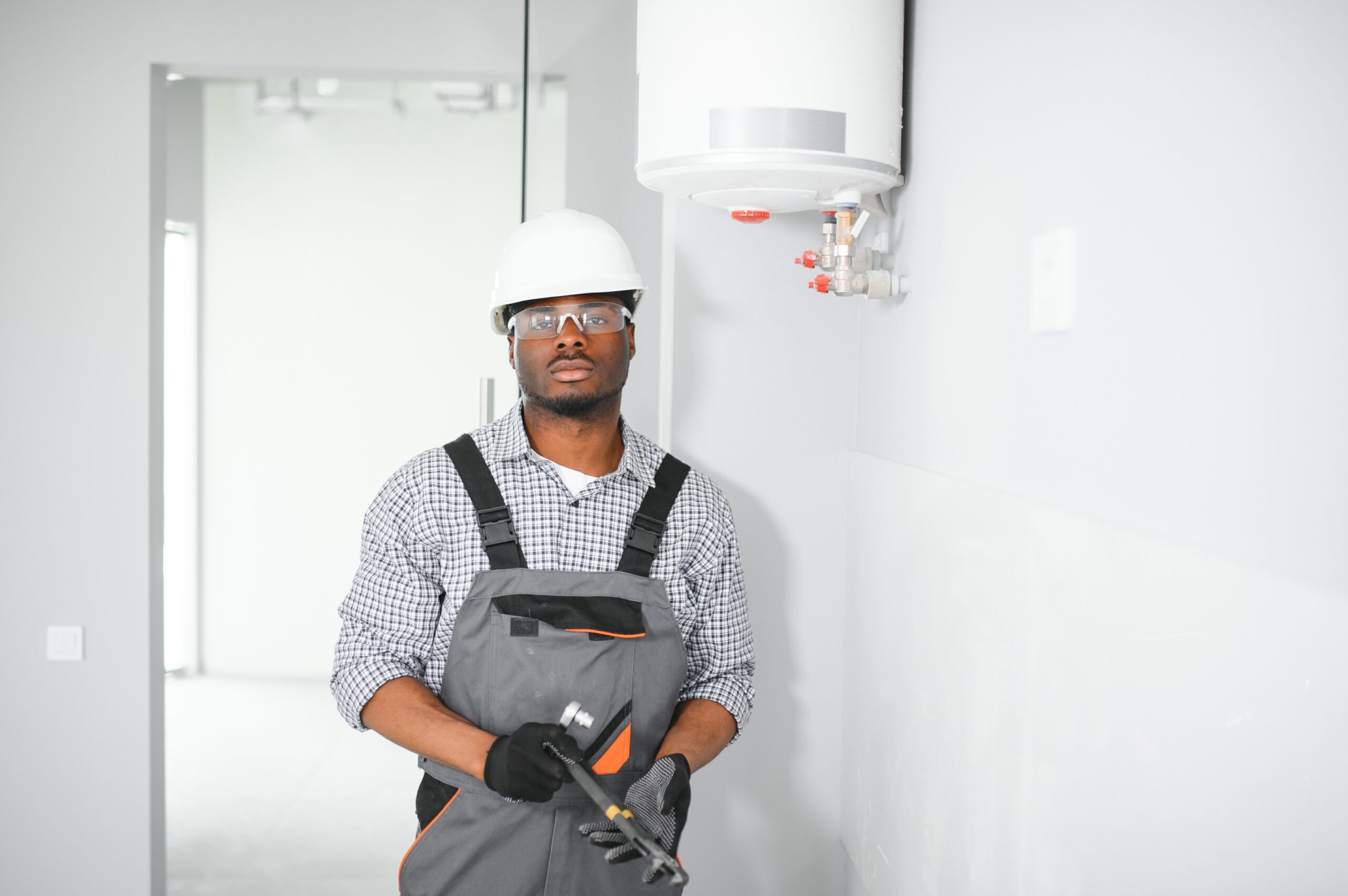 Plumbing Solutions LLC technician standing beside a wall-mounted residential water heater while holding maintenance tools.