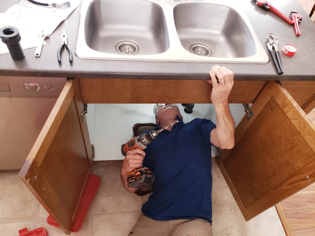 Professional plumber repairing kitchen plumbing under a sink using a power drill during leak inspection service.