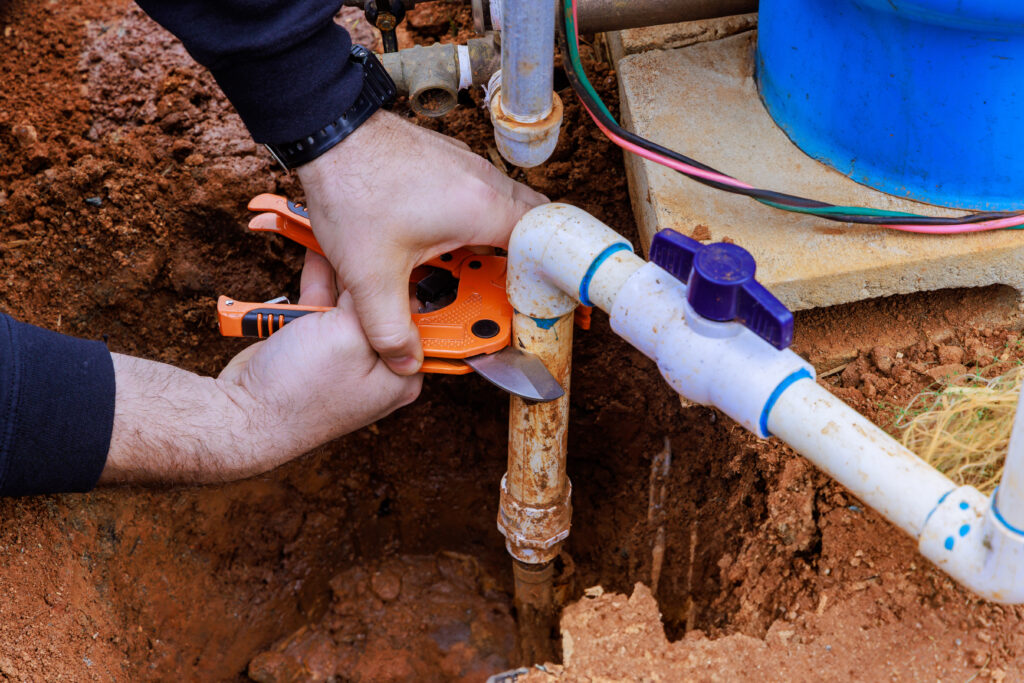 Technician cutting PVC water line in trench during outdoor plumbing and backflow installation work.