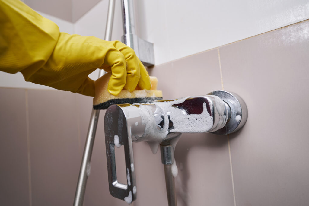 Gloved hand cleaning a chrome shower faucet with soap foam during bathroom fixture maintenance.