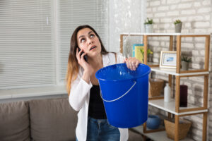Homeowner catching water dripping from a ceiling leak into a bucket while speaking on the phone inside a living room.