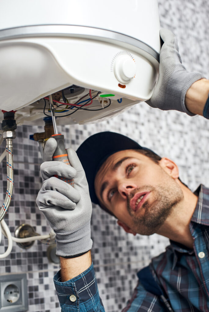 Plumber inspecting and repairing the underside connections of a residential water heater.