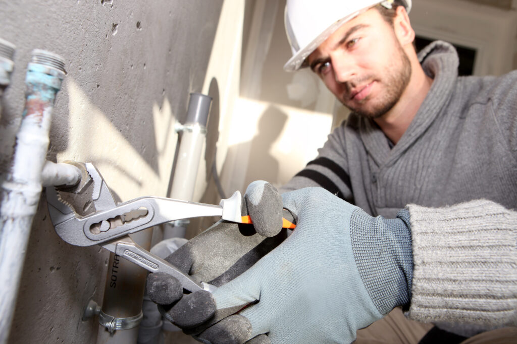 Plumbing Solutions LLC technician wearing a hard hat and gloves while repairing industrial plumbing pipes on-site.