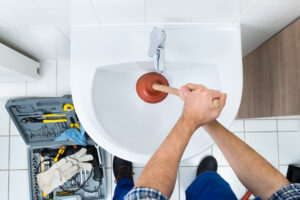 Plumbing Solutions LLC technician using a plunger to clear a clogged bathroom sink with plumbing tools arranged on the floor nearby.