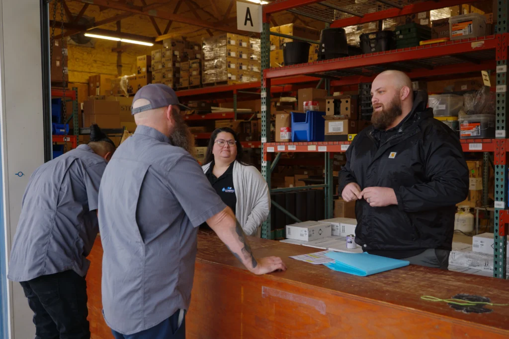 Plumbing Solutions LLC team members reviewing plumbing parts and supplies inside a stocked service warehouse.