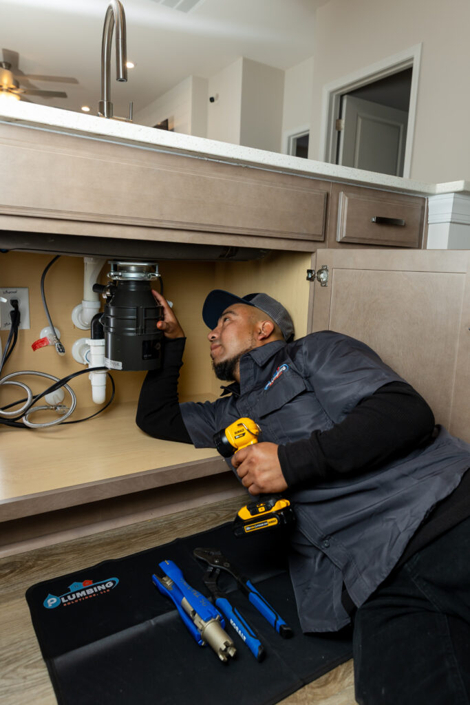 Plumbing Solutions LLC technician inspecting and servicing a garbage disposal beneath a kitchen sink to diagnose a recurring drain clog.