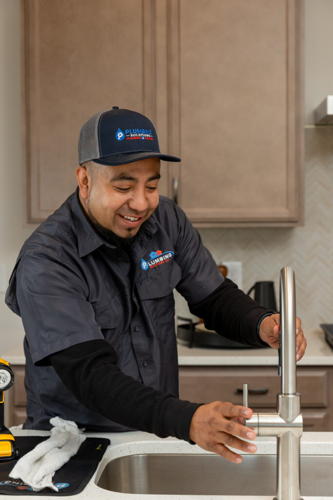 Plumbing Solutions LLC technician installing and connecting a kitchen sink faucet beneath a residential sink.